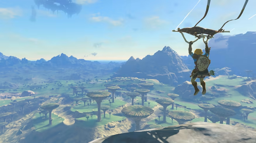 Link holds a kite-like glider overhead as he glides high above the land. Beneath him is a sprawling landscape with massive shroom like trees with flat tops, and mountain ranges in the distance.