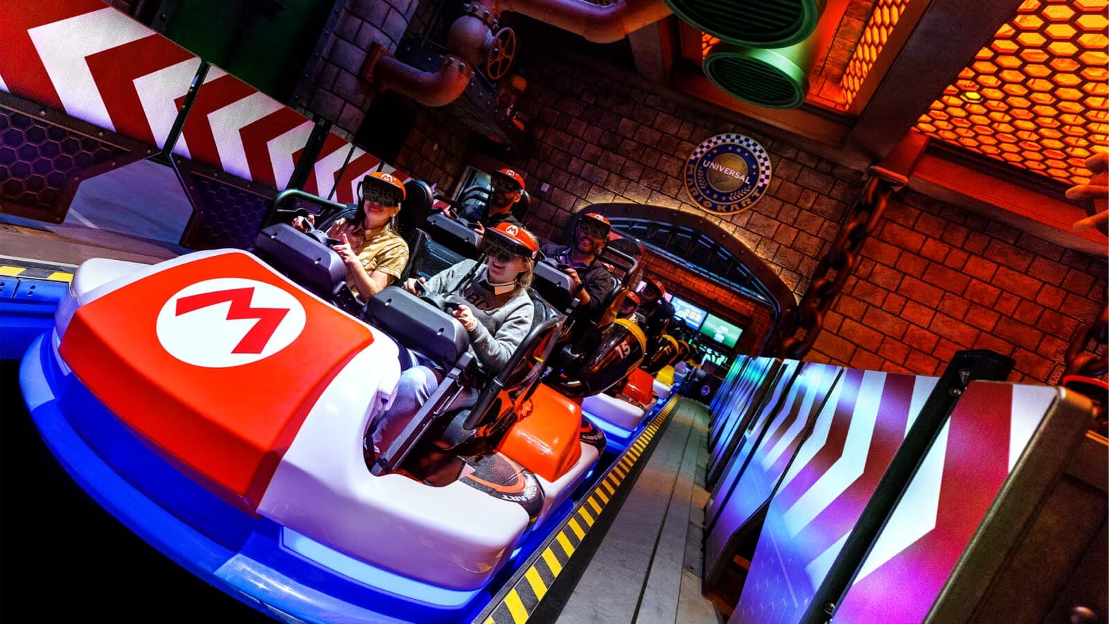 Families sitting in Mario Kart-like go karts ready to race in Mario Kart: Bowser's Challenge at Super Nintendo World in Universal Studios Hollywood. Attendees are seated four to a go-kart and are waring headsets as part of the ride. They are on a track that is similar to Bowser's Castle from Super Mario.