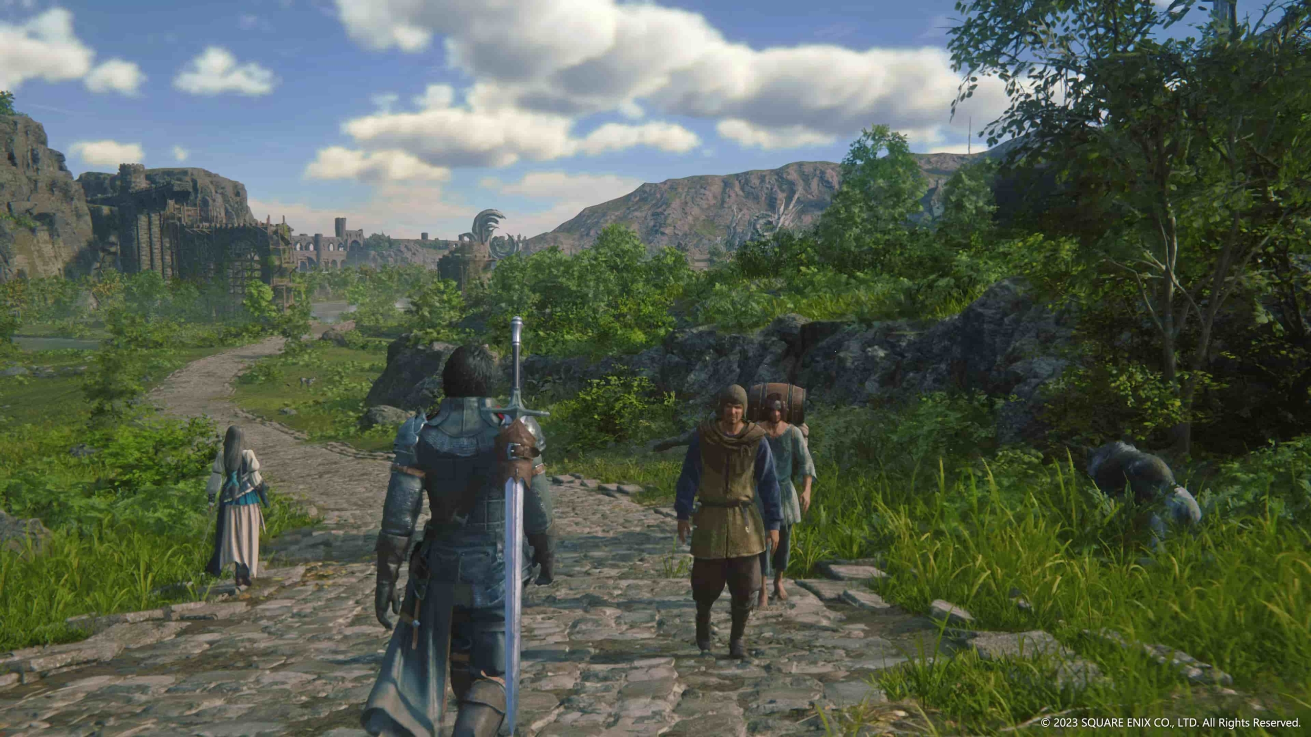 Clive stands on a medieval looking cobblestone road facing a vast landscape. The In the distance you can see gatehouse-like structures and mountains. The blue sky is dotted with puffy clouds.