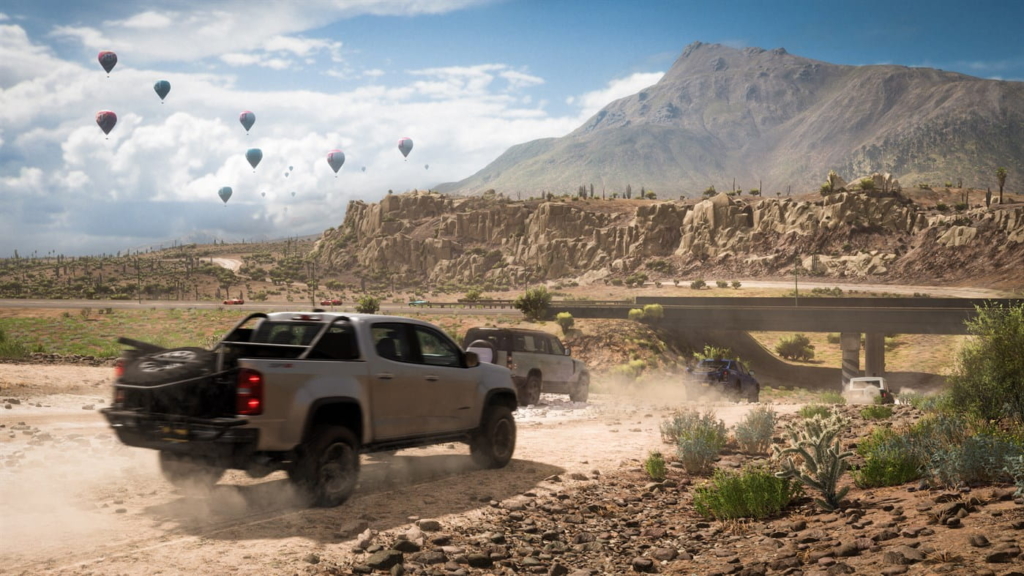 Several 4X4s speed across a dusty desert road in Forza Horizon 5. The sky in the distance is dotted with hot air balloons. Further in the distance, you can see hills and mountains peppered with desert shrubbery. 