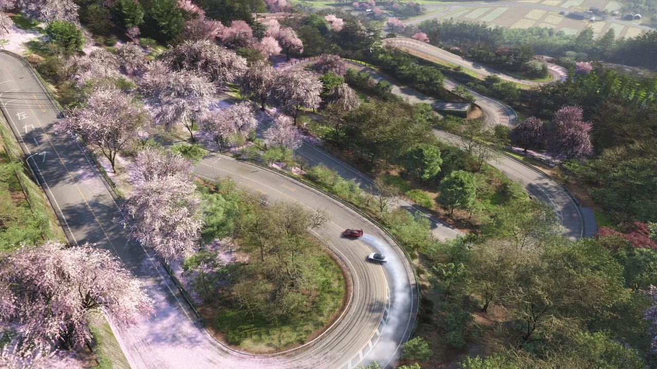 Two cars are seen drifting down a winding road in the Japanese countryside. Cherry blossom trees and other lush greenery are scattered around the perimeter of the roads.